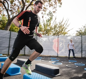 A man is actively participating in an outdoor fitness session, jumping over blue platforms with agility. Two other people in the background are also engaging in exercise, with rings scattered on the ground. The area is fenced and surrounded by trees, suggesting a park or recreational setting.